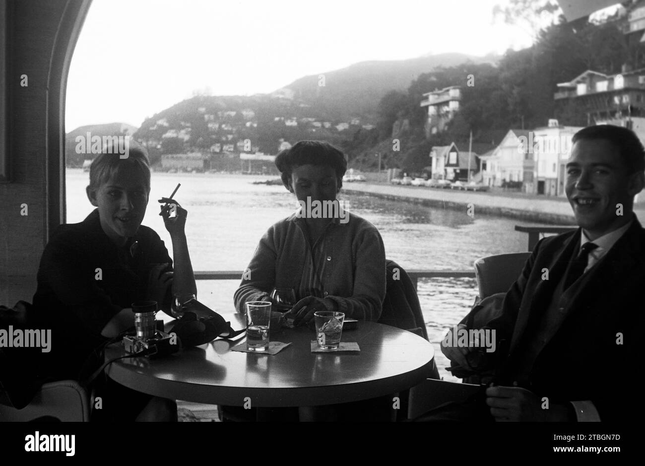 Zwei Frauen und ein Mann BEI Nachmittagsdrinks in einem Lokal am Wasser, eine Kamera liegt auf dem Tisch vor ihnen, San Francisco 1962. Deux femmes et un homme prenant un verre après-midi dans un pub au bord de l'eau, une caméra couchée sur la table en face d'eux, San Francisco 1962. Banque D'Images