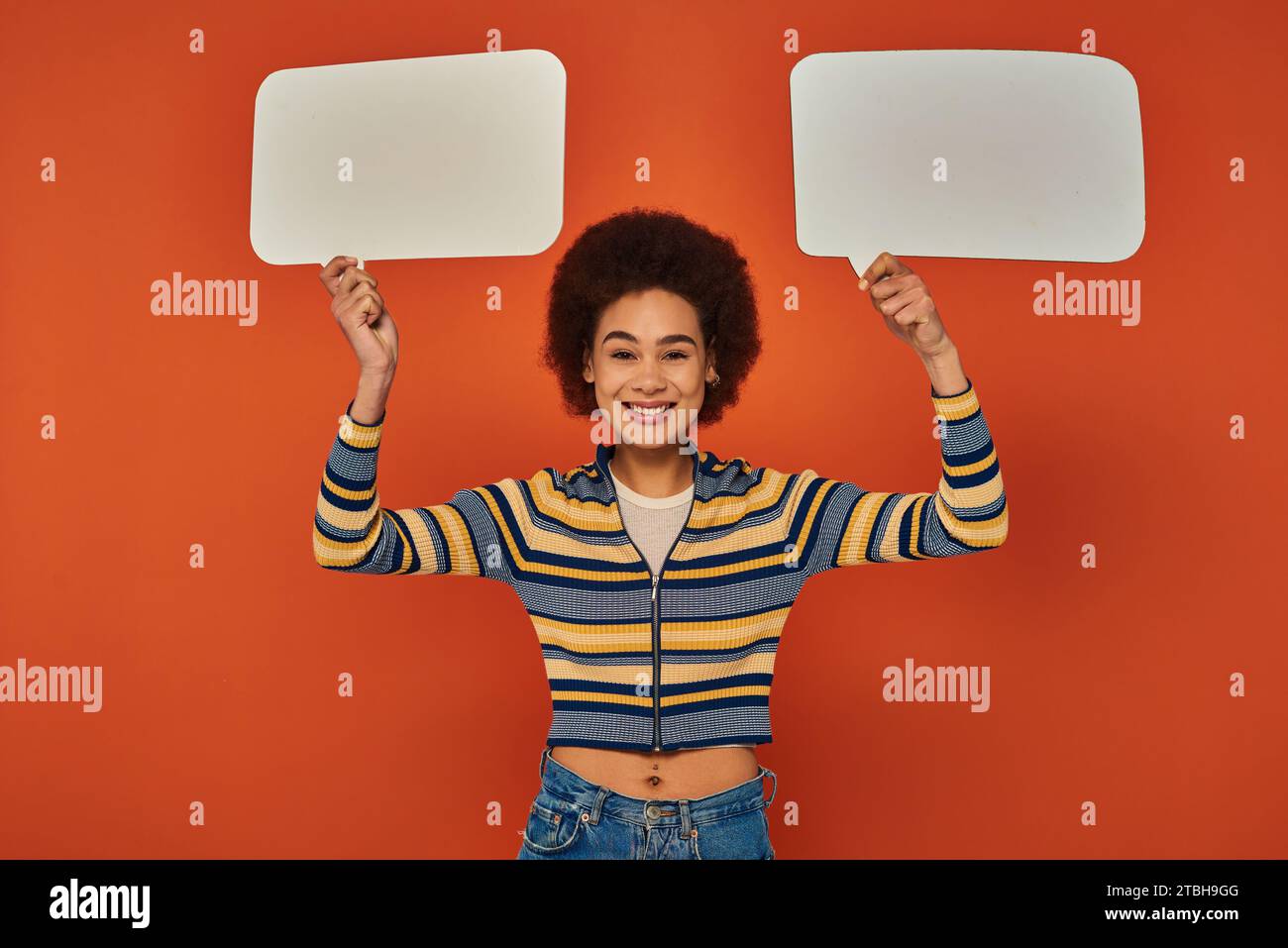 joyeuse femme afro-américaine en tenue vibrante posant avec des bulles de parole et souriant à la caméra Banque D'Images
