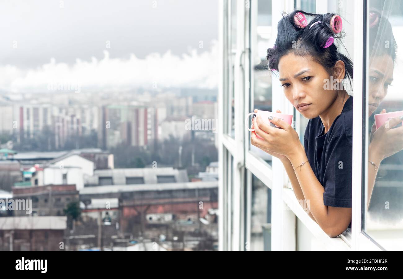 Une jeune femme en curleurs regarde par la fenêtre d'un immeuble de grande hauteur dans une zone industrielle Banque D'Images