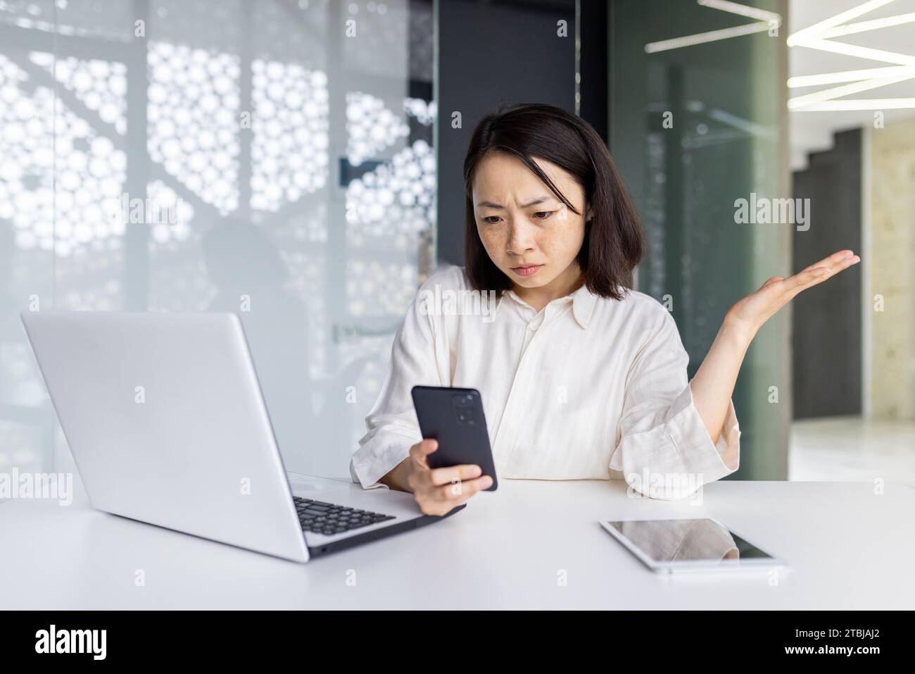Femme triste bouleversée au lieu de travail tenant un smartphone, femme d'affaires déprimée naviguant sur les médias sociaux à l'intérieur du bureau, travailleuse asiatique utilisant un ordinateur portable au travail. Banque D'Images