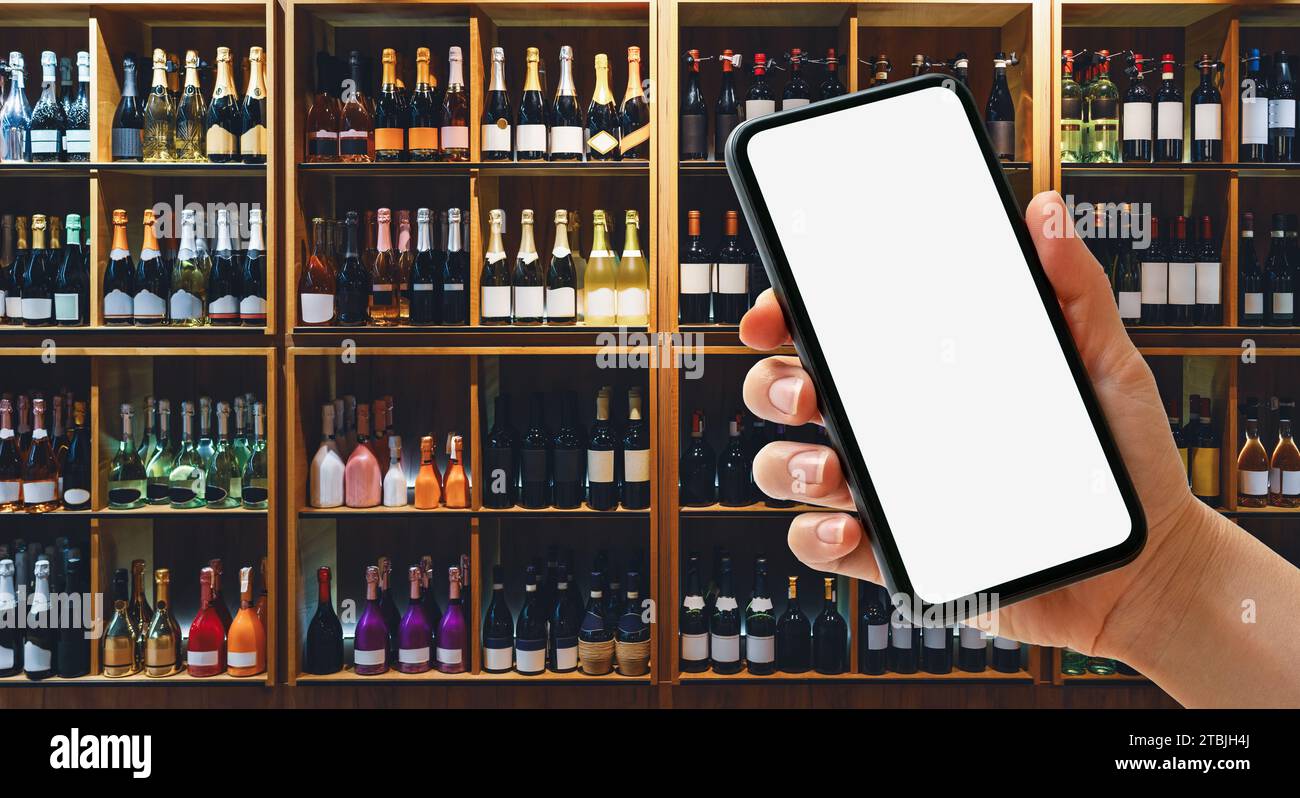 Téléphone intelligent avec écran blanc dans la main féminine devant les étagères avec des bouteilles de vin dans le magasin d'alcool. Banque D'Images