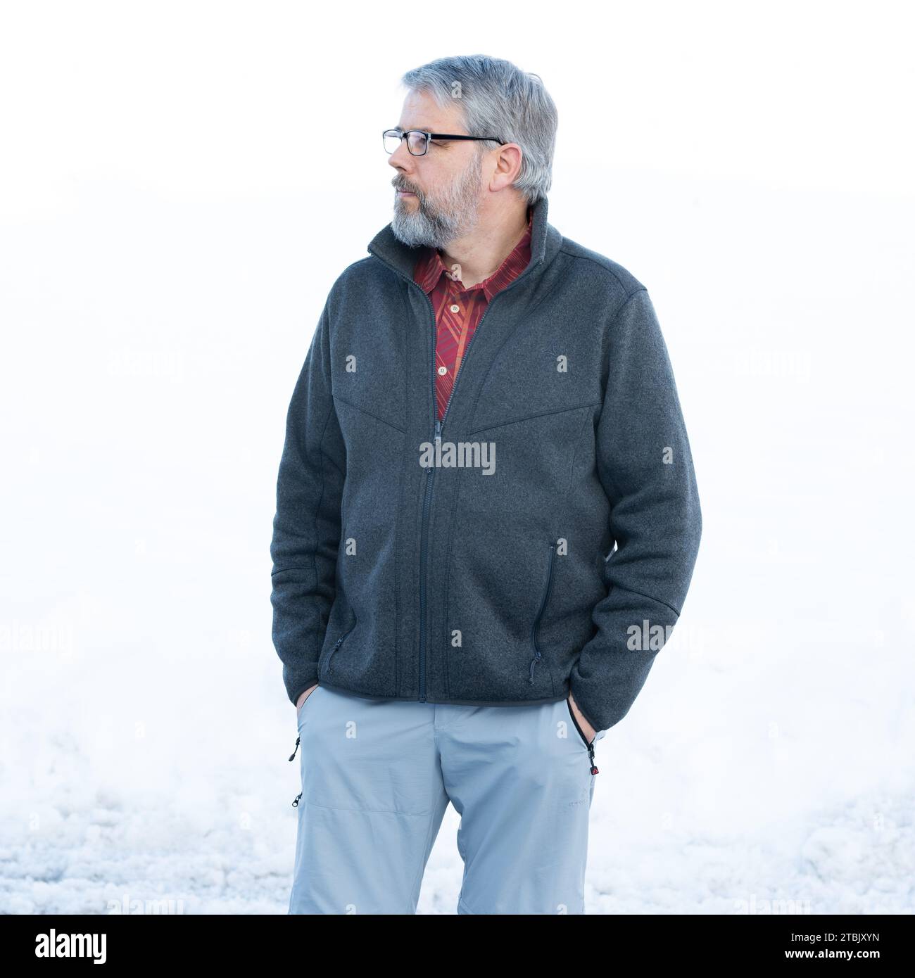 Homme, 56 ans, cheveux gris et barbe, debout dehors en hiver avec de la neige en arrière-plan, les mains en poche, portant des lunettes Banque D'Images