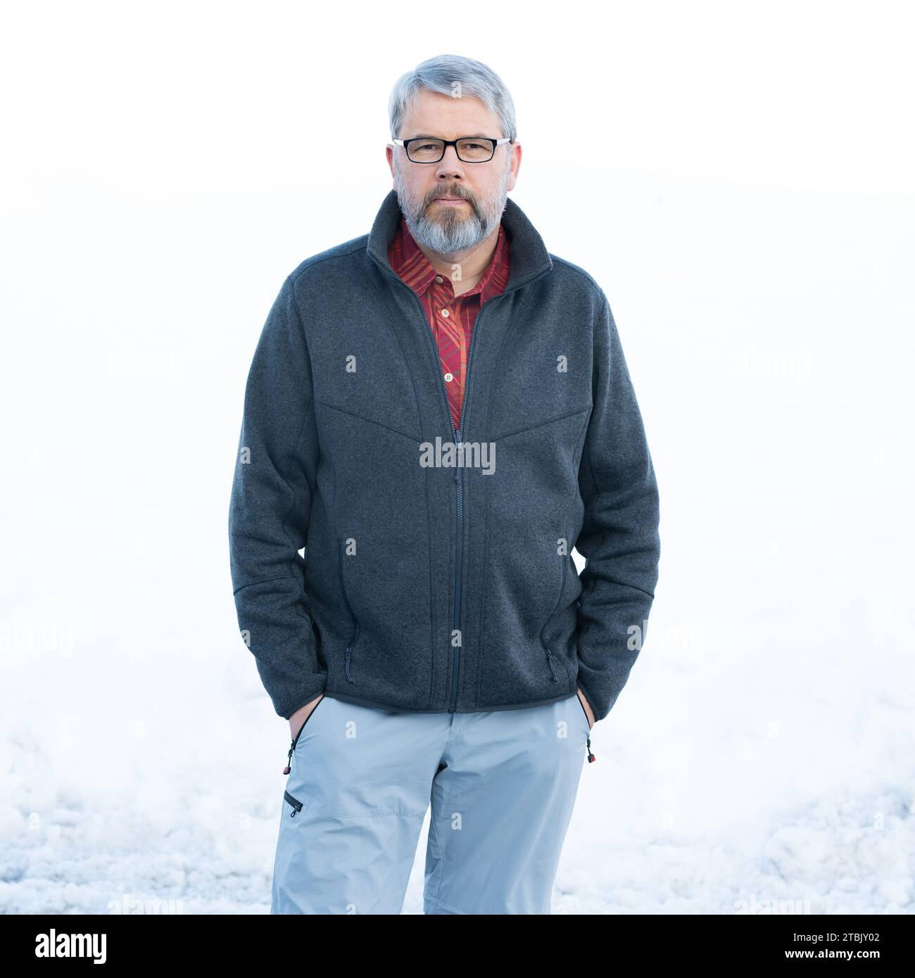 Homme, 56 ans, cheveux gris et barbe, debout dehors en hiver avec de la neige en arrière-plan, les mains en poche, portant des lunettes Banque D'Images