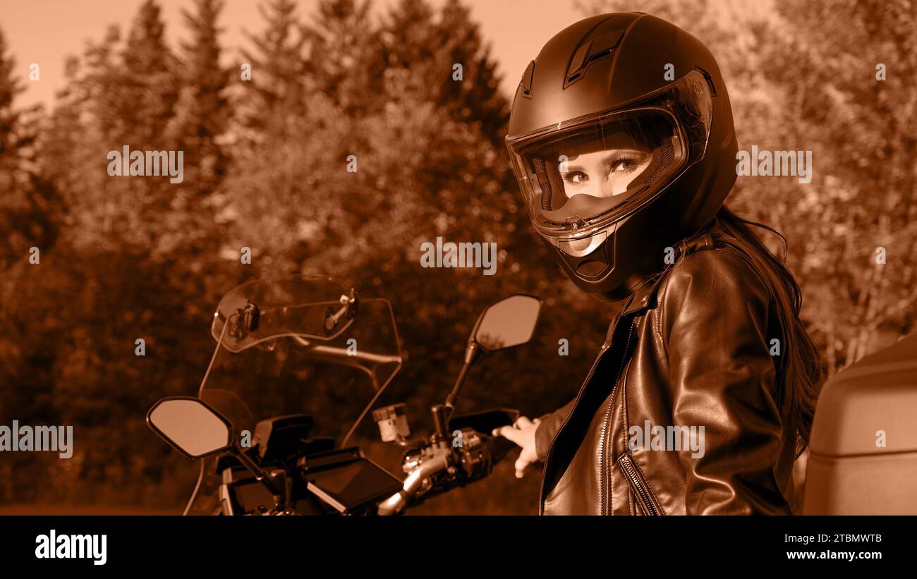 Moto girl chevauchant la moto de tourisme sur la route forestière. Image teintée dans la couleur Peach Fuzz de l'année 2024. Belle femme avec les cheveux rouges dans la veste en cuir Banque D'Images