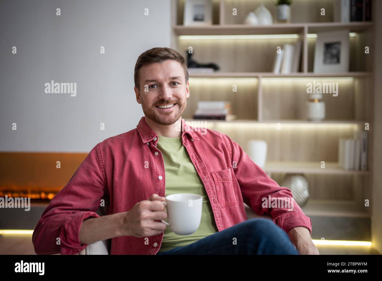 Heureux homme souriant amical assis dans un appartement confortable avec tasse de thé, boire, se reposer, regarder la caméra Banque D'Images