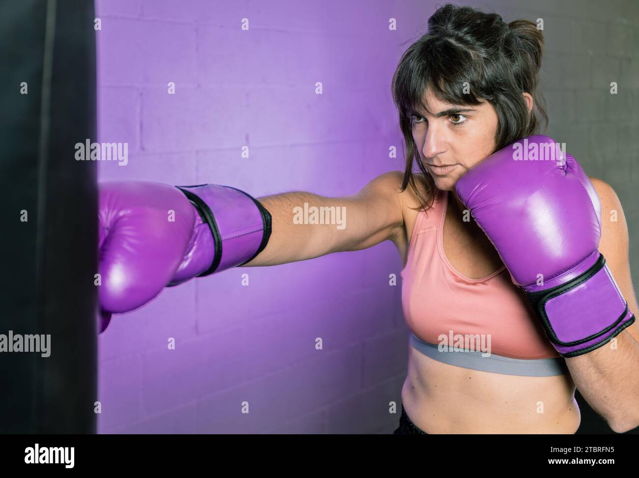 Photo horizontale femmes mi adulte boxe caucasienne avec sac et gants violets dans la salle de gym. Sur fond d'espace de copie violet. Féminisme. Sports, loisirs Banque D'Images