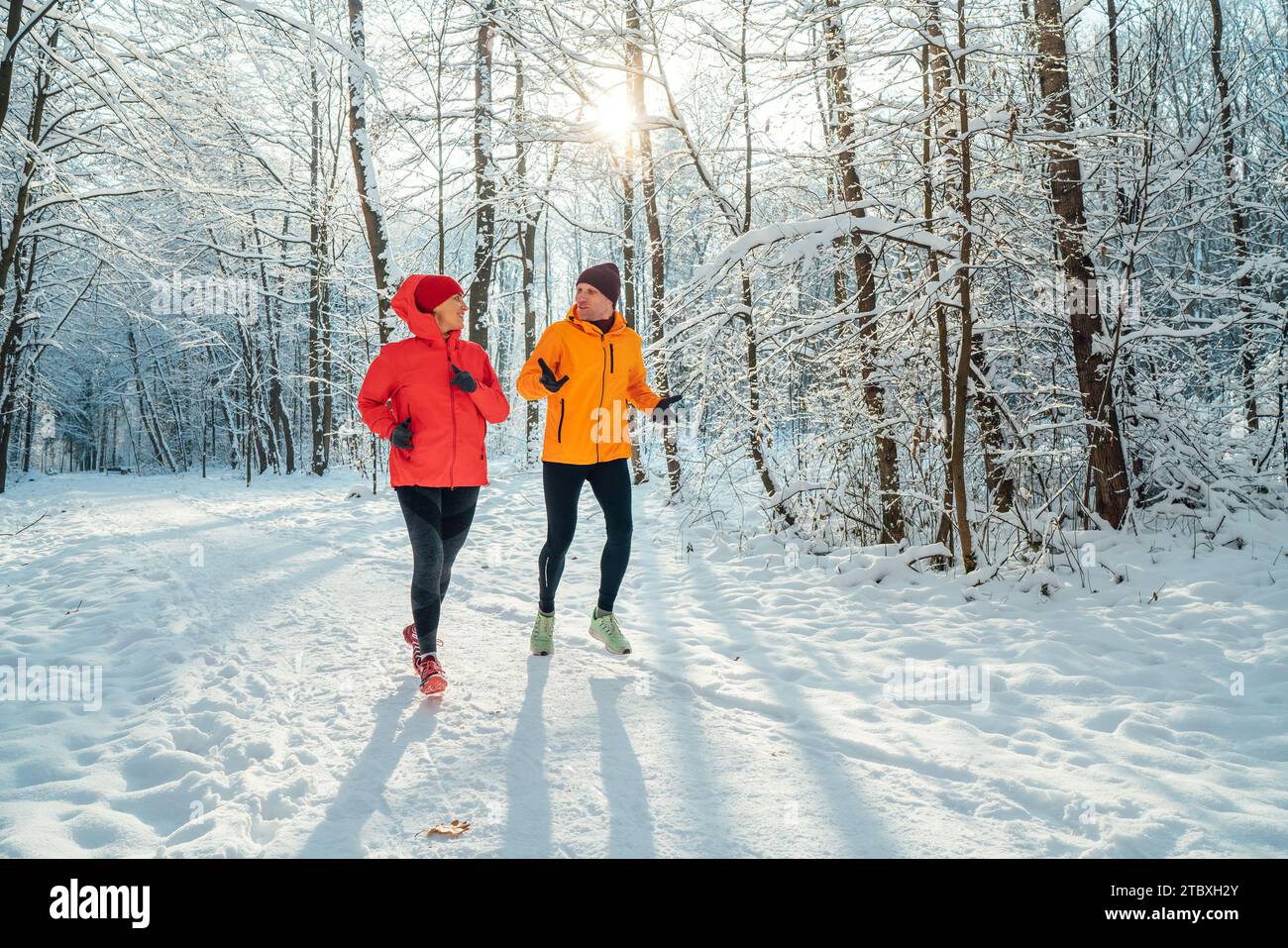 Un couple de coureurs souriants d'âge moyen homme et femme habillés de vestes coupe-vent brillantes courant parlant forêt enneigée pittoresque pendant le soleil gelé Banque D'Images