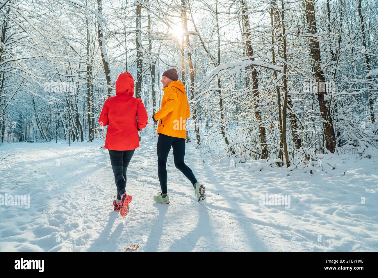 Un couple de coureurs souriants d'âge moyen homme et femme habillés de vestes coupe-vent brillantes courant parlant forêt enneigée pittoresque pendant le soleil gelé Banque D'Images