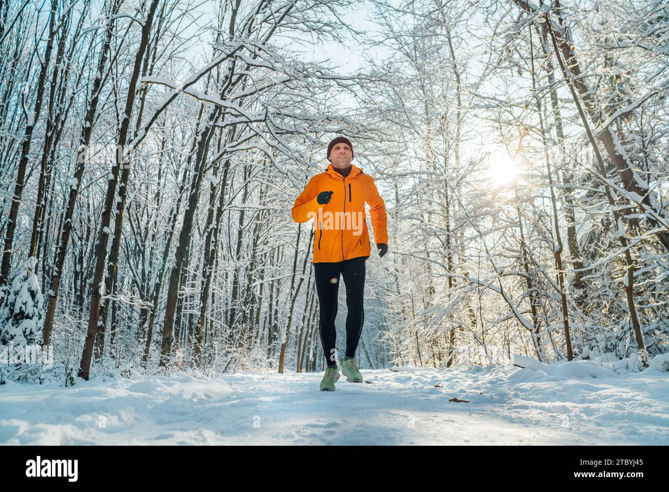 Homme de coureur de Trail souriant d'âge moyen vêtu d'une veste coupe-vent orange vif endurance course rapide forêt enneigée pittoresque pendant une journée ensoleillée gelée. S Banque D'Images