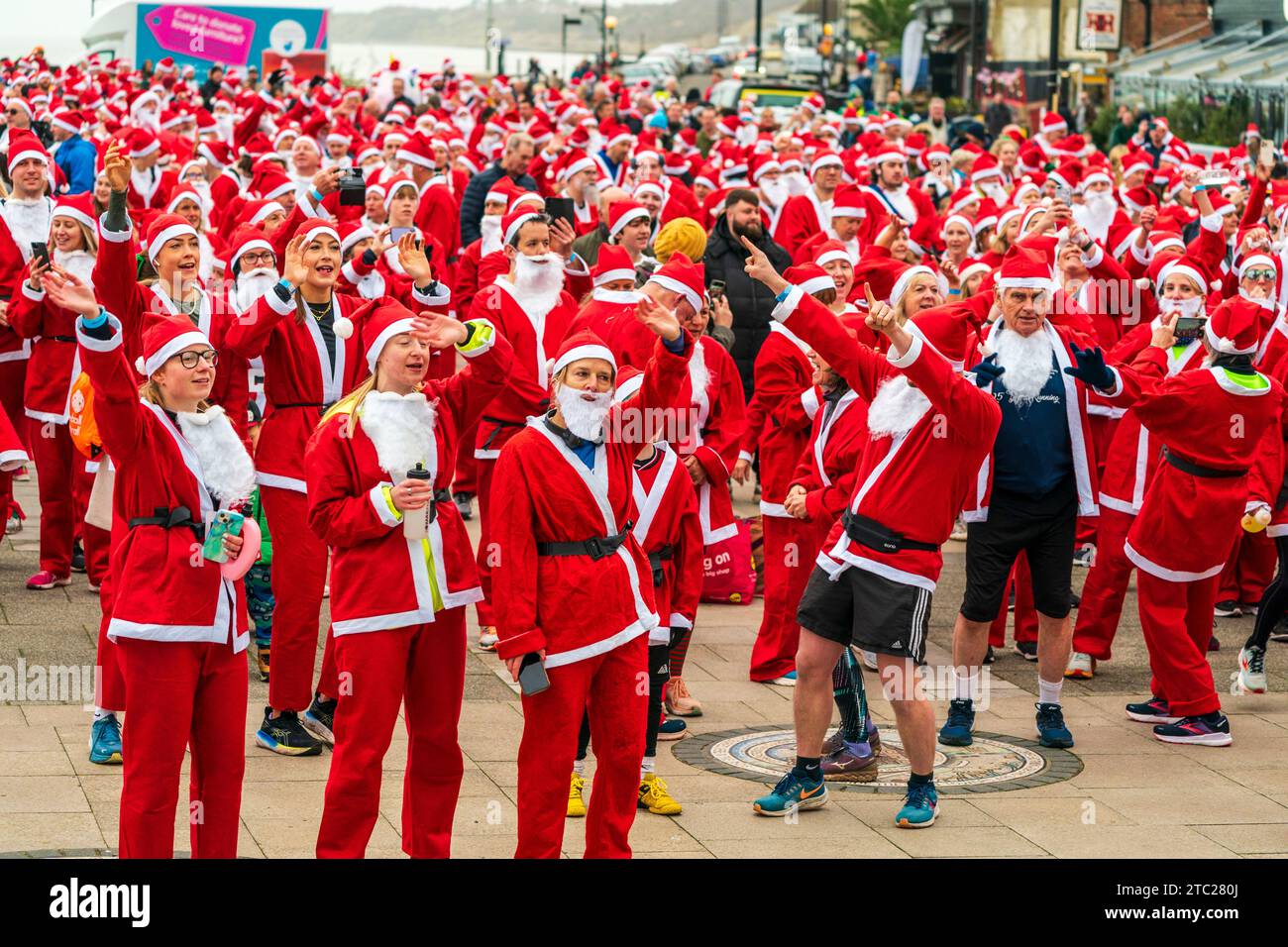 Plusieurs centaines de personnes toutes vêtues de vêtements rouges du Père Noël debout en plein air sur le front de mer à Herne Bay avant de prendre part à une course caritative, 'Santa's in the Bay' pour le Pilgrims Hospice. la plupart d'entre eux acclament et les bras levés. Banque D'Images