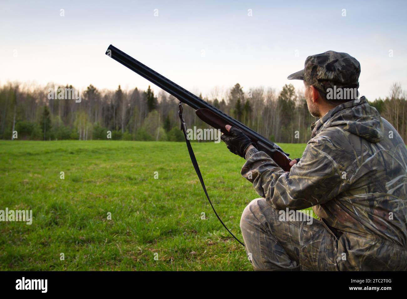 un chasseur avec un fusil de chasse dans ses mains s'agenouilla et regarde une clairière dans la soirée dans la forêt printanière Banque D'Images