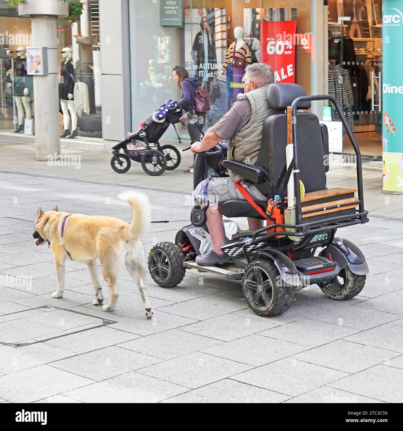 Homme conduisant un scooter pour personnes handicapées dans un centre commercial piétonnier emmenant son chien pour une promenade sur une laisse attenante à Bury St Edmunds Suffolk Angleterre Royaume-Uni Banque D'Images