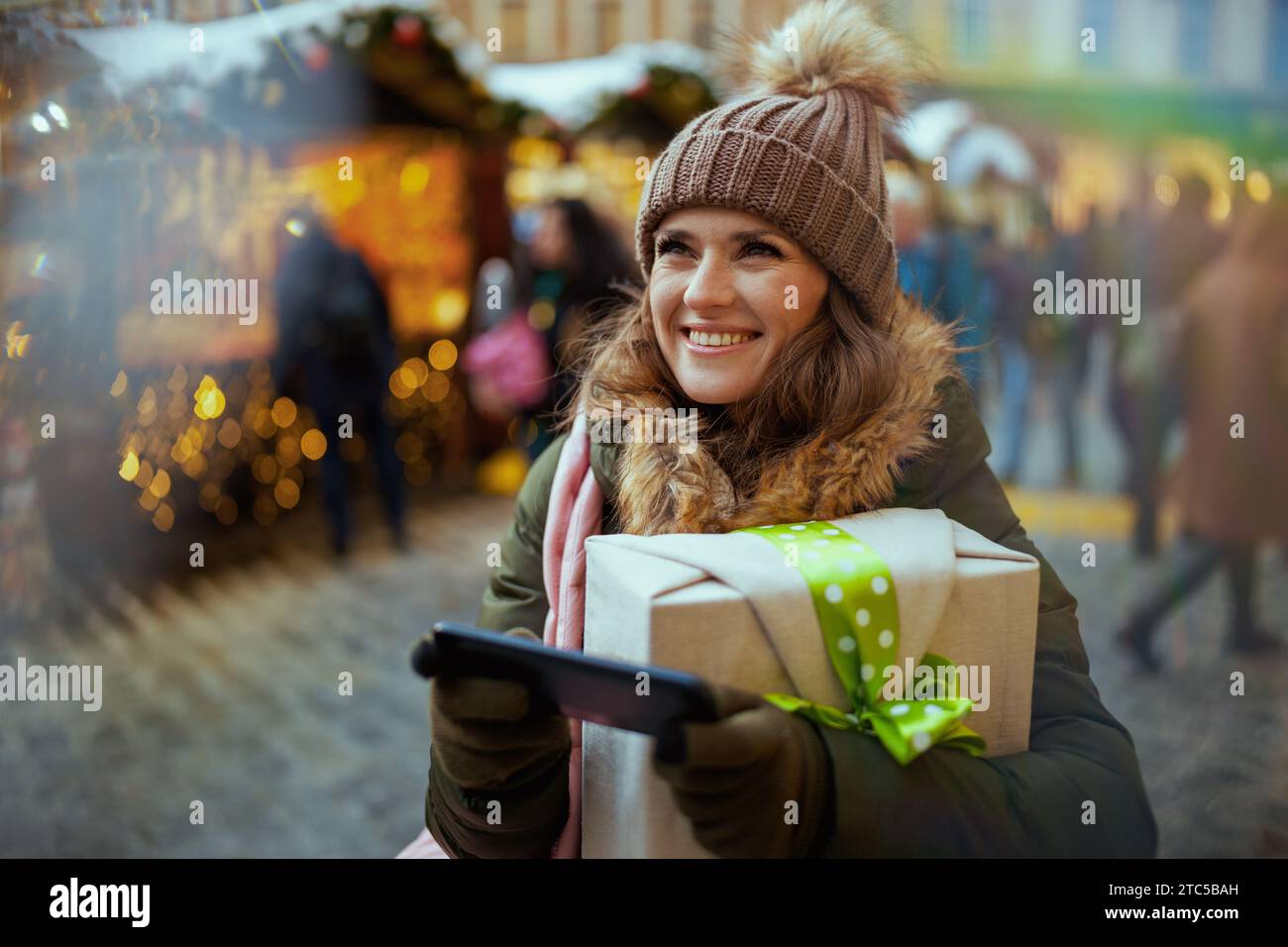 heureuse femme moderne en manteau vert et chapeau brun à la foire de noël dans la ville en utilisant smartphone. Banque D'Images