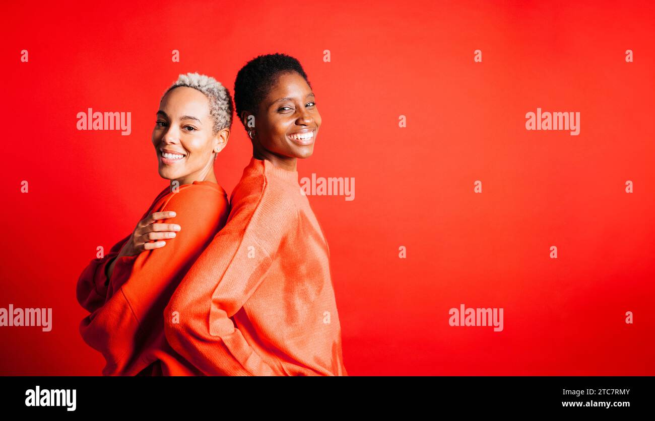 Portrait de deux jeunes couples de femmes, portant un pull rouge, embrassant devant un fond rouge au studio. Deux femmes ensemble au studio. Banque D'Images