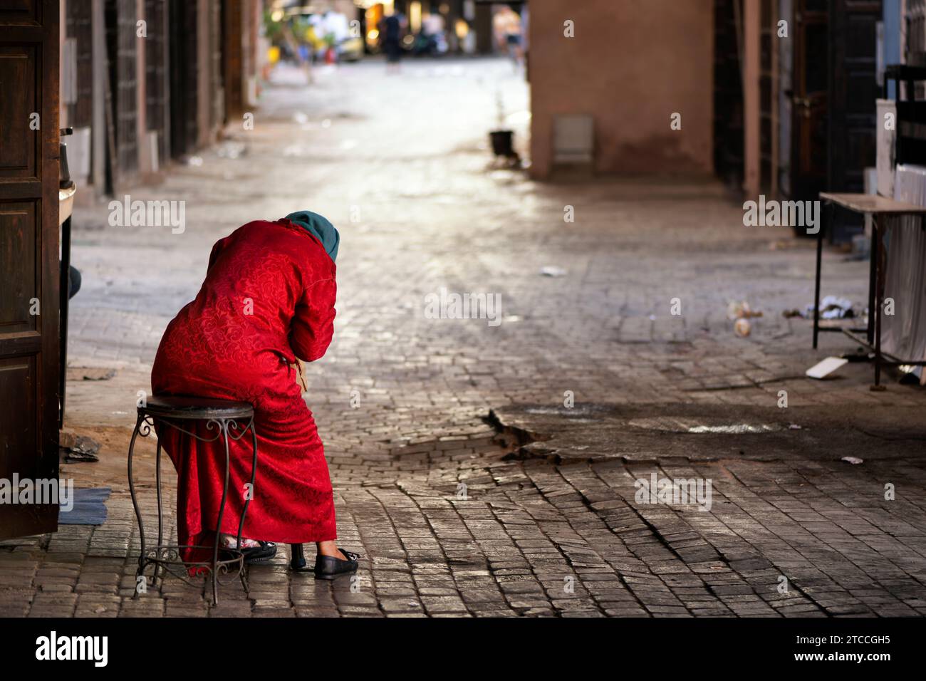 Femme âgée marocaine fatiguée habillée rouge prenant un repos sur un tabouret placé sur une ruelle à l'intérieur du souk de Marrakech ou Médina de Marrakech. Banque D'Images