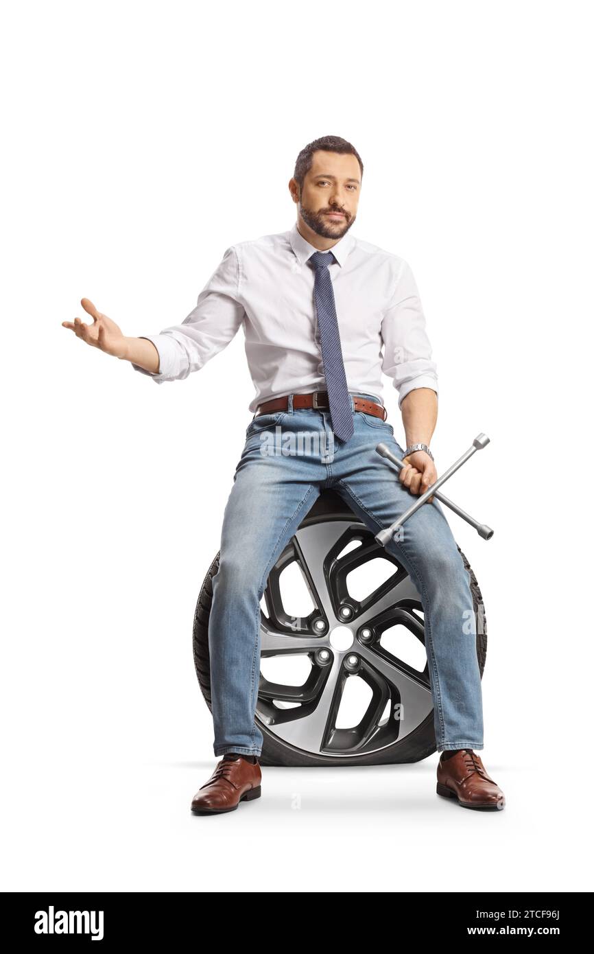 Homme sérieux assis sur un pneu de voiture à plat et tenant une clé à écrous isolé sur fond blanc Banque D'Images