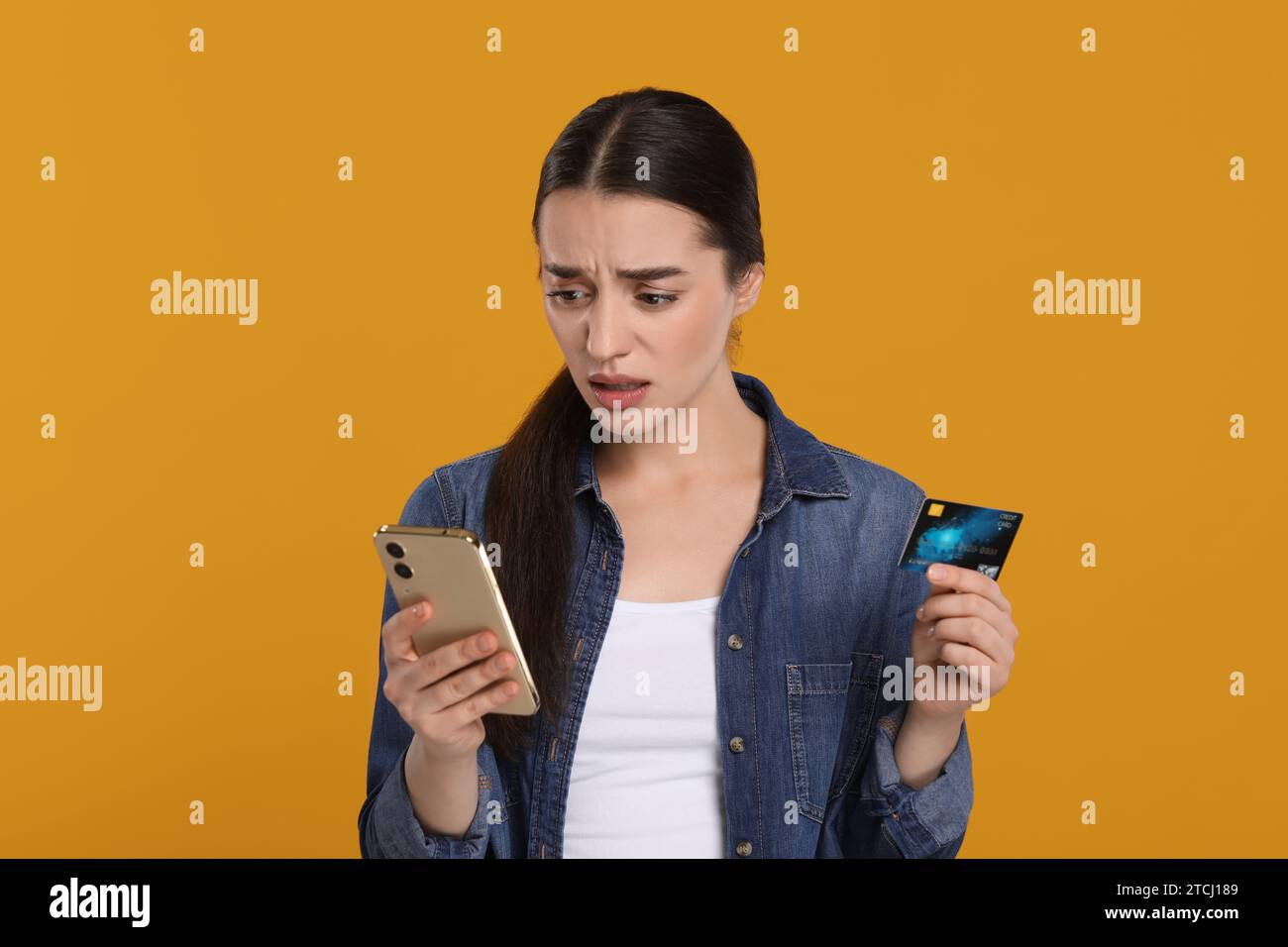Femme confuse avec carte de crédit et smartphone sur fond orange. Problème de dette Banque D'Images