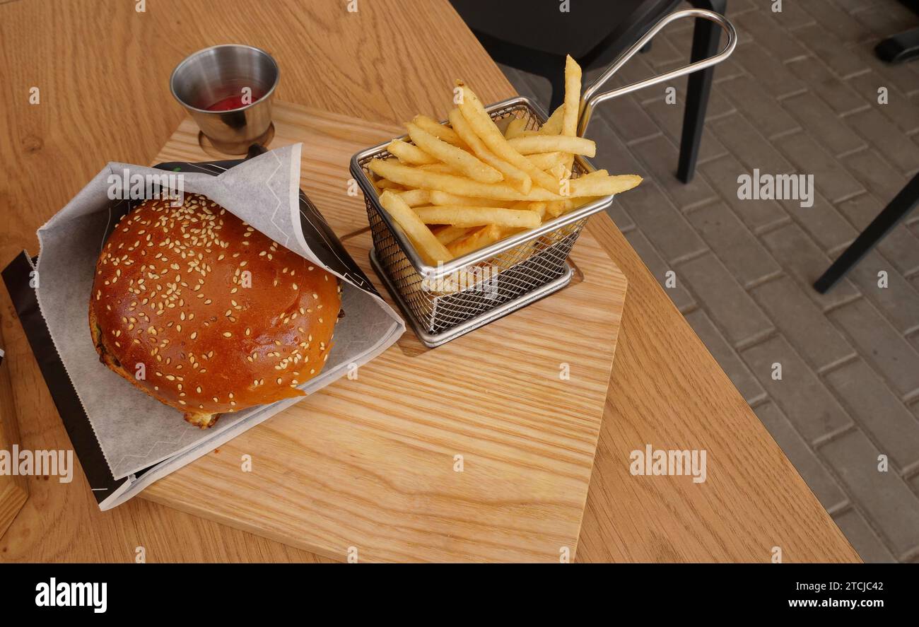 Hamburger dans du papier d'emballage et frites dans un panier en maille métallique sur une table au snack-bar en plein air Banque D'Images