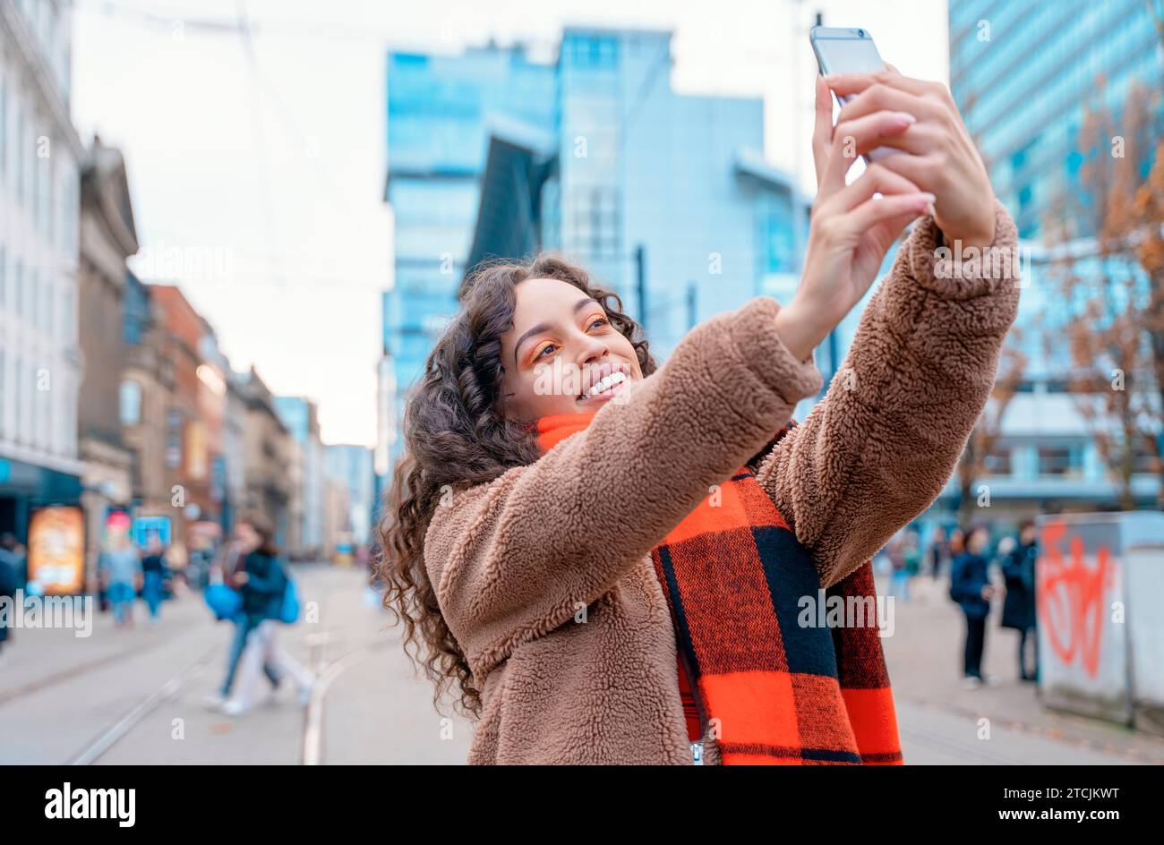Une femme gaie qui prend des selfies sur un smartphone Une dame brune dans une veste et une écharpe orange souriant et voyageant en Europe. Banque D'Images