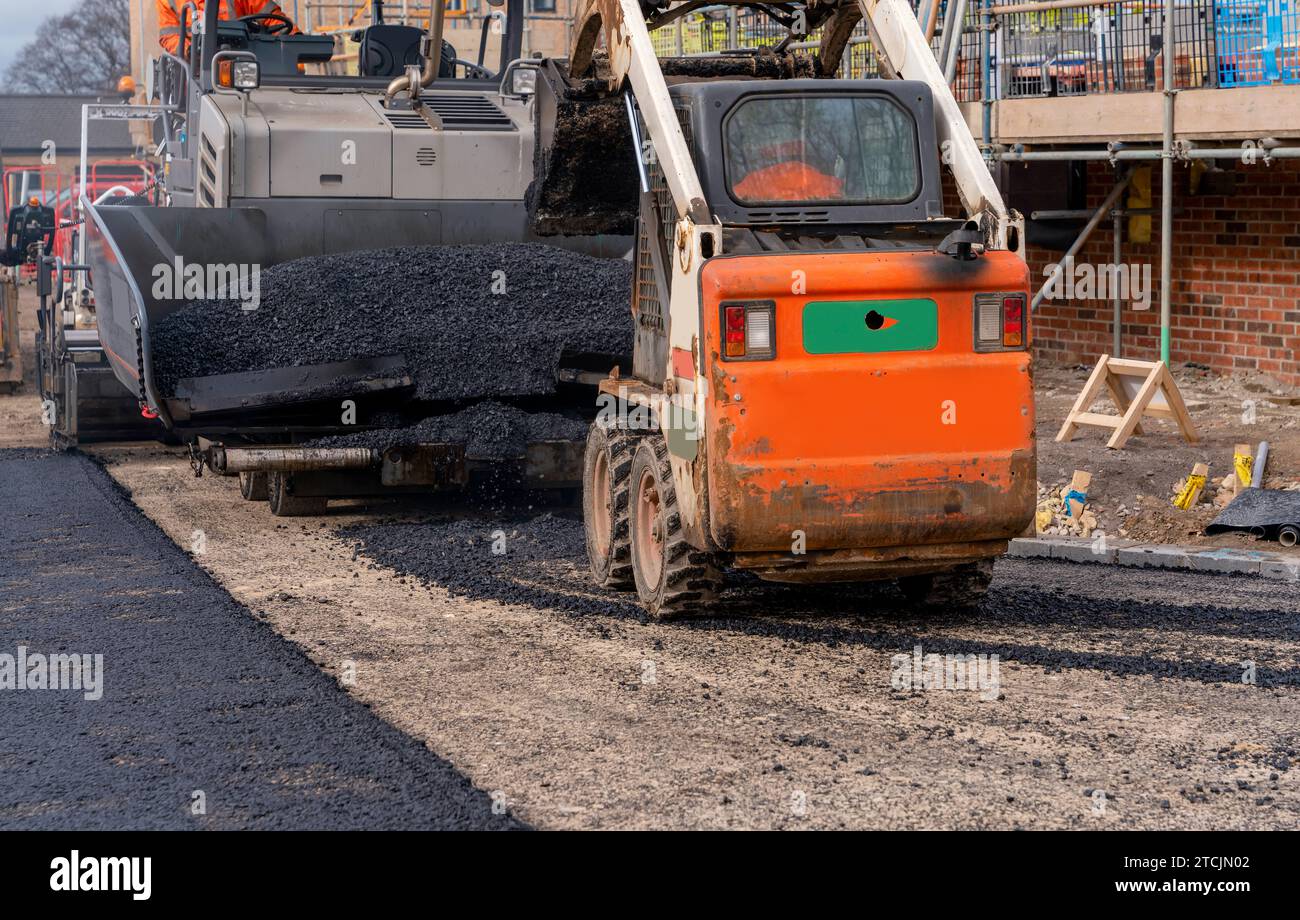 Finisseur d'asphalte rempli de tarmac chaud posant la nouvelle surface de route sur le nouveau site de développement de logements résidentiels et opérateur routier en orange hi-viz Next Banque D'Images