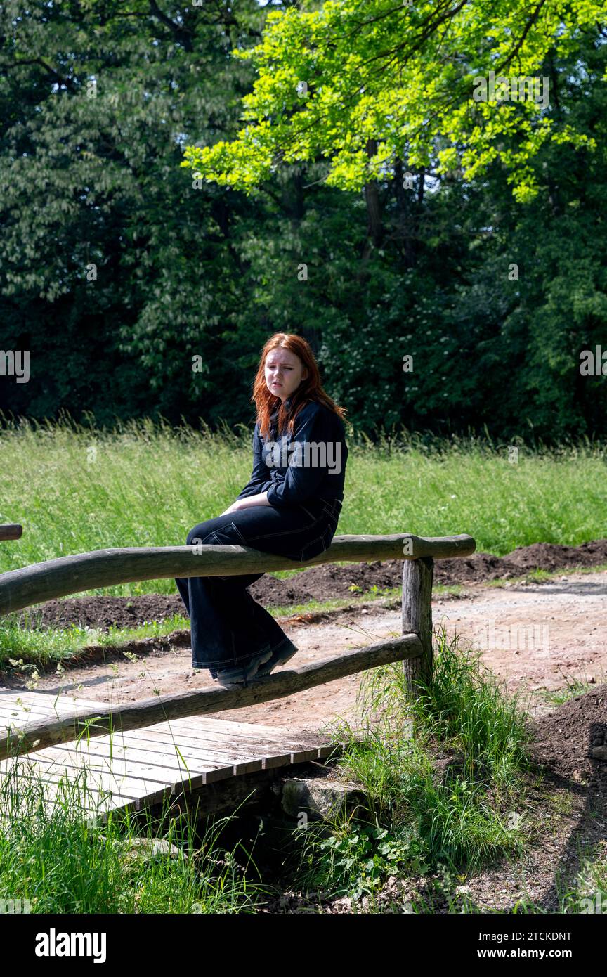 Une adolescente aux cheveux roux est assise tristement sur la balustrade en bois d'un pont dans la nature verte par une journée d'été ensoleillée Banque D'Images
