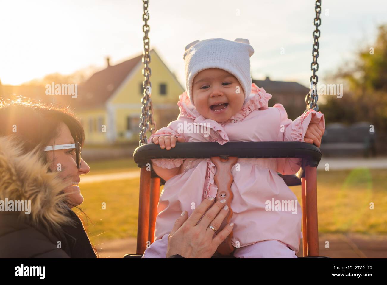 Adorable baby girl avec de grands beaux yeux et un bonnet s'amusant sur une balançoire ride à une aire de jeux dans un parc ensoleillé Banque D'Images