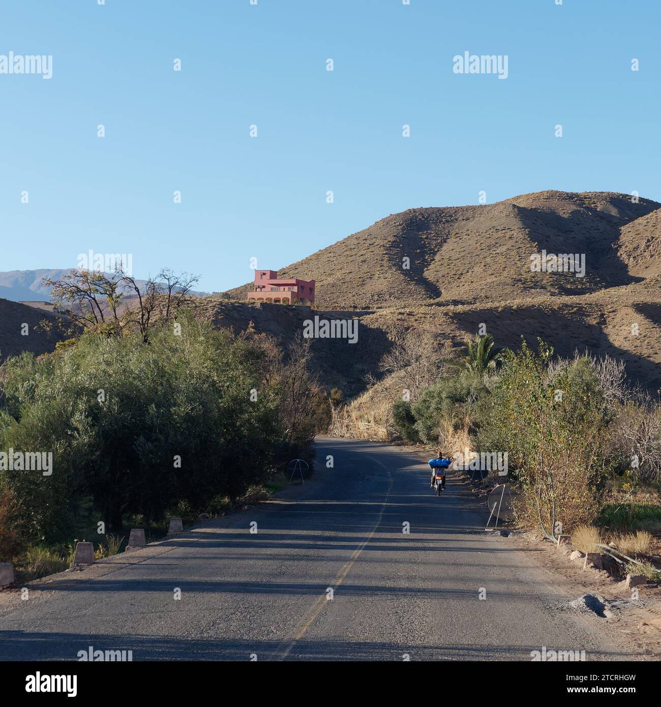 Route bordée d'arbres avec moto et maison sur une colline parmi les montagnes de l'Atlas à l'est de Marrakech, Maroc, 13 décembre 2023 Banque D'Images