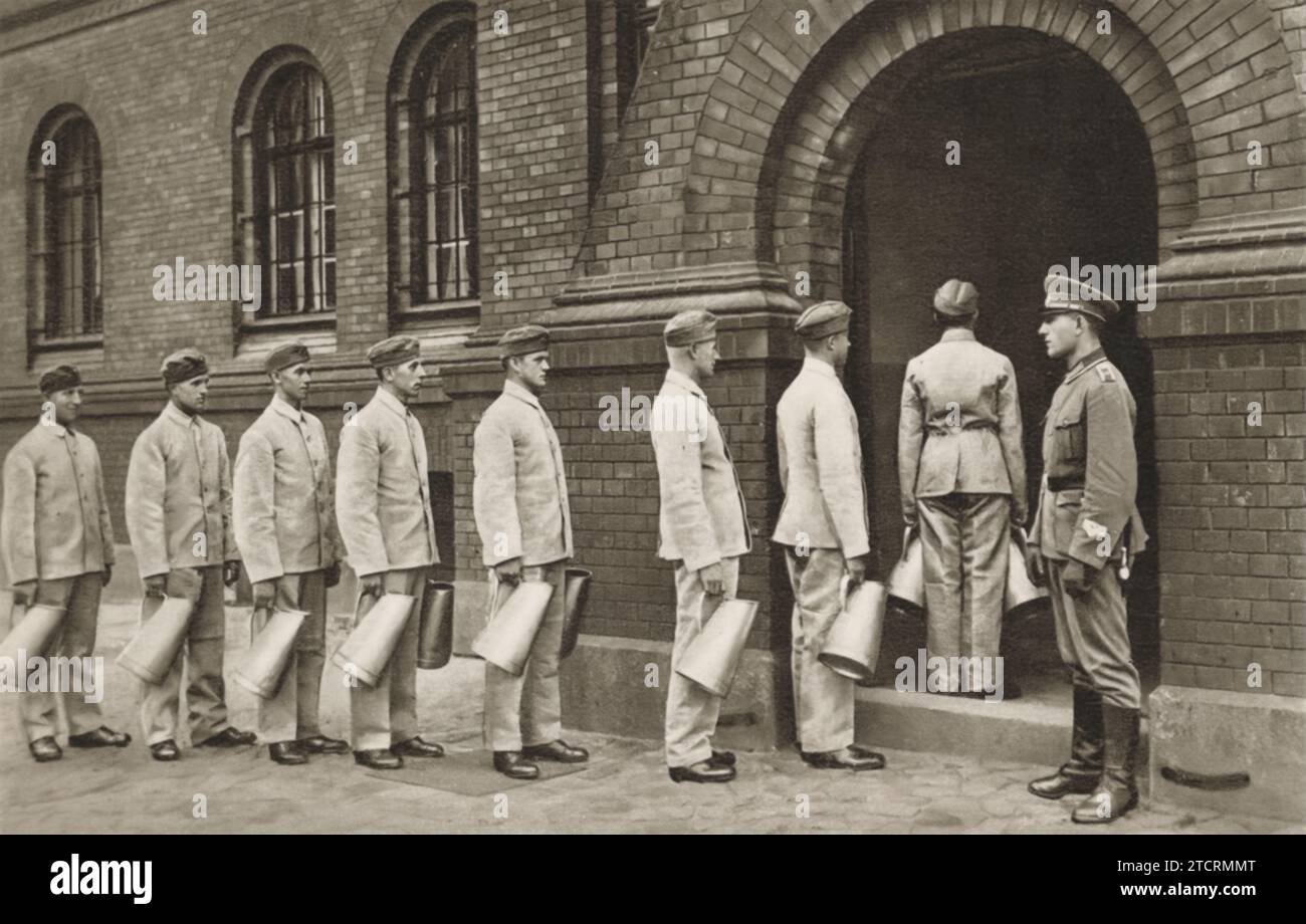 Les soldats allemands sont alignés à l'extérieur de la cuisine à 5:15 heures du matin, à peine 15 minutes après leur premier appel de réveil. Probablement désignés comme « passeurs de café », leur tâche est de recueillir et de distribuer le café à leurs camarades soldats. La scène reflète l'importance de tâches, même petites, dans le maintien du moral et de la cohésion de l'unité, en commençant la journée avec une dose vitale d'énergie et de travail d'équipe. Banque D'Images