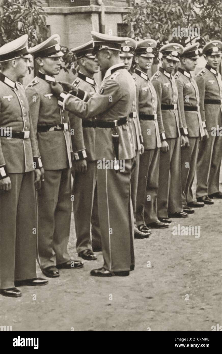 Un officier supérieur est vu corriger méticuleusement l'uniforme d'un soldat allemand, suggérant la préparation d'un événement important, probablement une inspection finale avant un défilé ou peut-être leur diplôme de formation militaire. Ce moment souligne l’attention au détail et à la précision attendues dans l’apparence militaire, en particulier pour les cérémonies importantes. L ' inspection reflète à la fois la discipline inculquée aux soldats et la fierté associée à leurs réalisations, marquant un point charnière dans leur voyage militaire. Banque D'Images