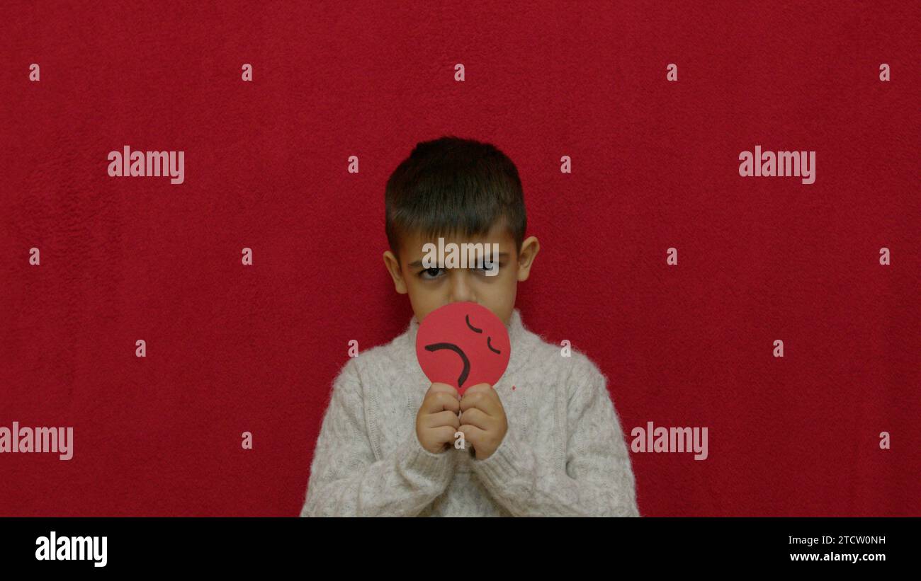 Un garçon en colère, un enfant exprimant des émotions, tenant un triste emoji rouge. Isolé sur rouge avec espace de copie. Photo de haute qualité Banque D'Images