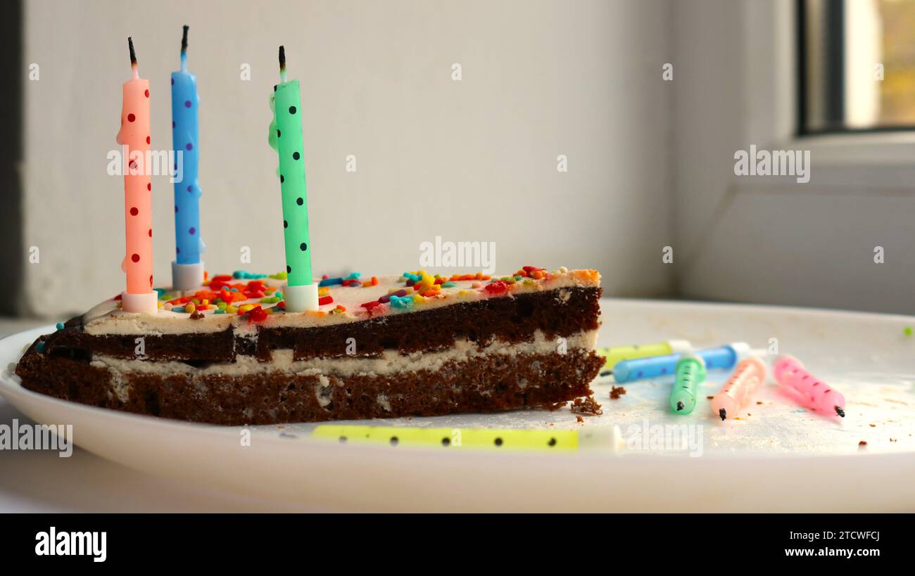 un morceau de gâteau fait de gâteau éponge au chocolat et de crème de lait blanc avec plusieurs bougies multicolores éteintes sur une assiette blanche comme concept Banque D'Images