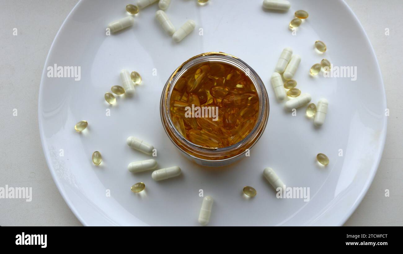 Pot de capsules d'huile de poisson doré avec des vitamines E et des graisses saines vue de dessus sur une grande assiette blanche avec d'autres capsules dispersées, prenant oméga 3 diététique Banque D'Images