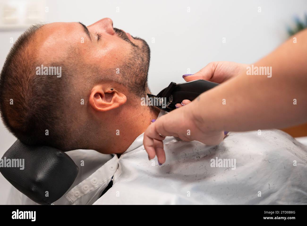 Vue latérale des mains d'un barbier coupé et méconnaissable rasant la barbe d'un client à l'œil fermé avec une tondeuse à gazon au salon Banque D'Images