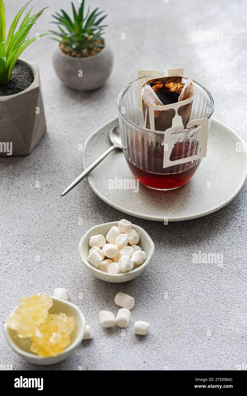Une tasse fraîche de café goutte à goutte dans un verre transparent avec fond gris tendance, accompagné de guimauves sucrées et de plantes décoratives chics. Banque D'Images