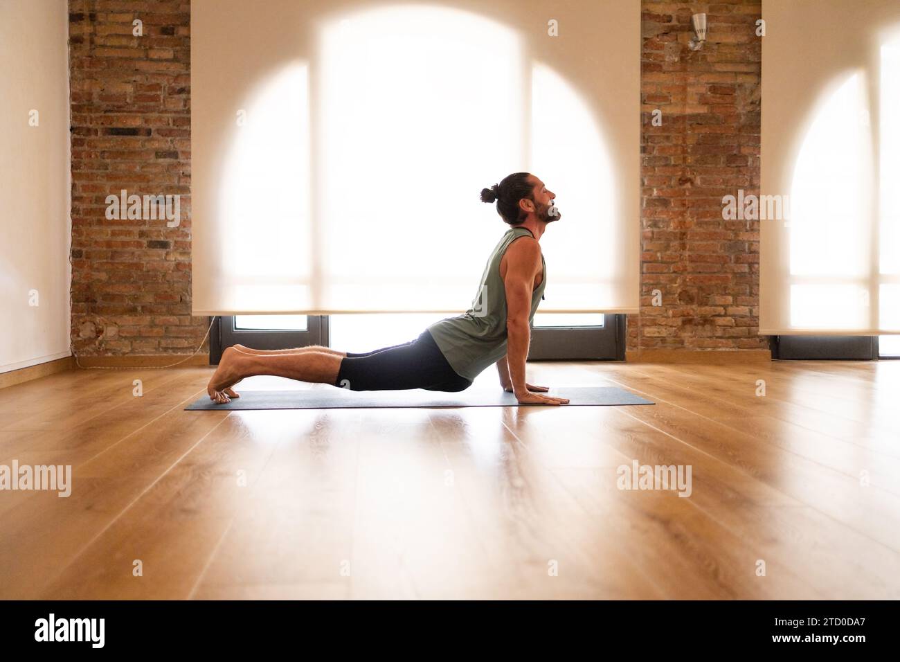 Un homme effectue un étirement de yoga sur un tapis dans une pièce avec des planchers de bois franc et des murs de briques apparentes, illuminés par la lumière naturelle. Banque D'Images