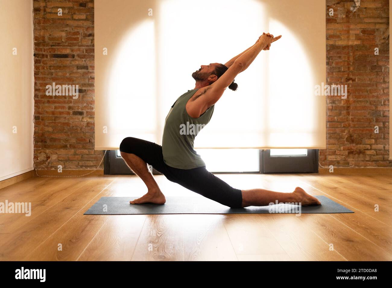 Un homme effectue un étirement de yoga sur un tapis dans une pièce avec des planchers de bois franc et des murs de briques apparentes, illuminés par la lumière naturelle. Banque D'Images