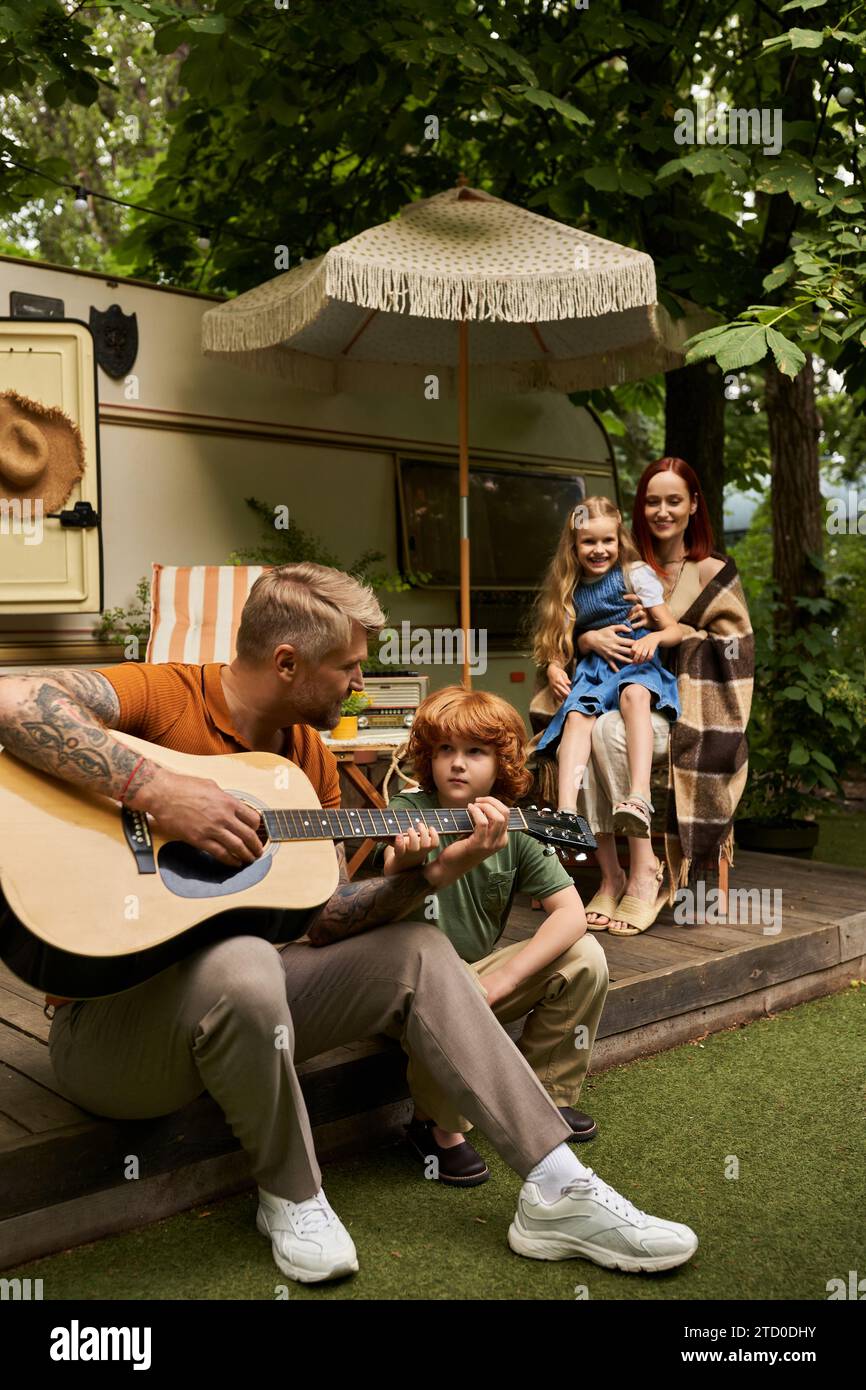 homme tatoué jouant de la guitare à un fils rousse attentif et la famille souriant à côté de la maison de remorque Banque D'Images