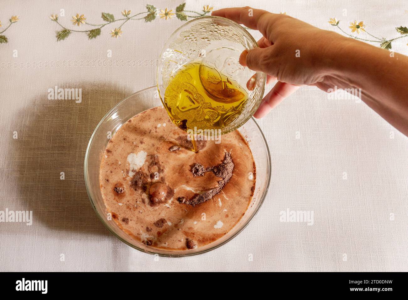 Vue de dessus de la personne anonyme de la récolte est montrée aspergeant d'huile d'olive sur un bol en verre clair rempli de lait et de chocolat en poudre, sur une table avec emboide Banque D'Images