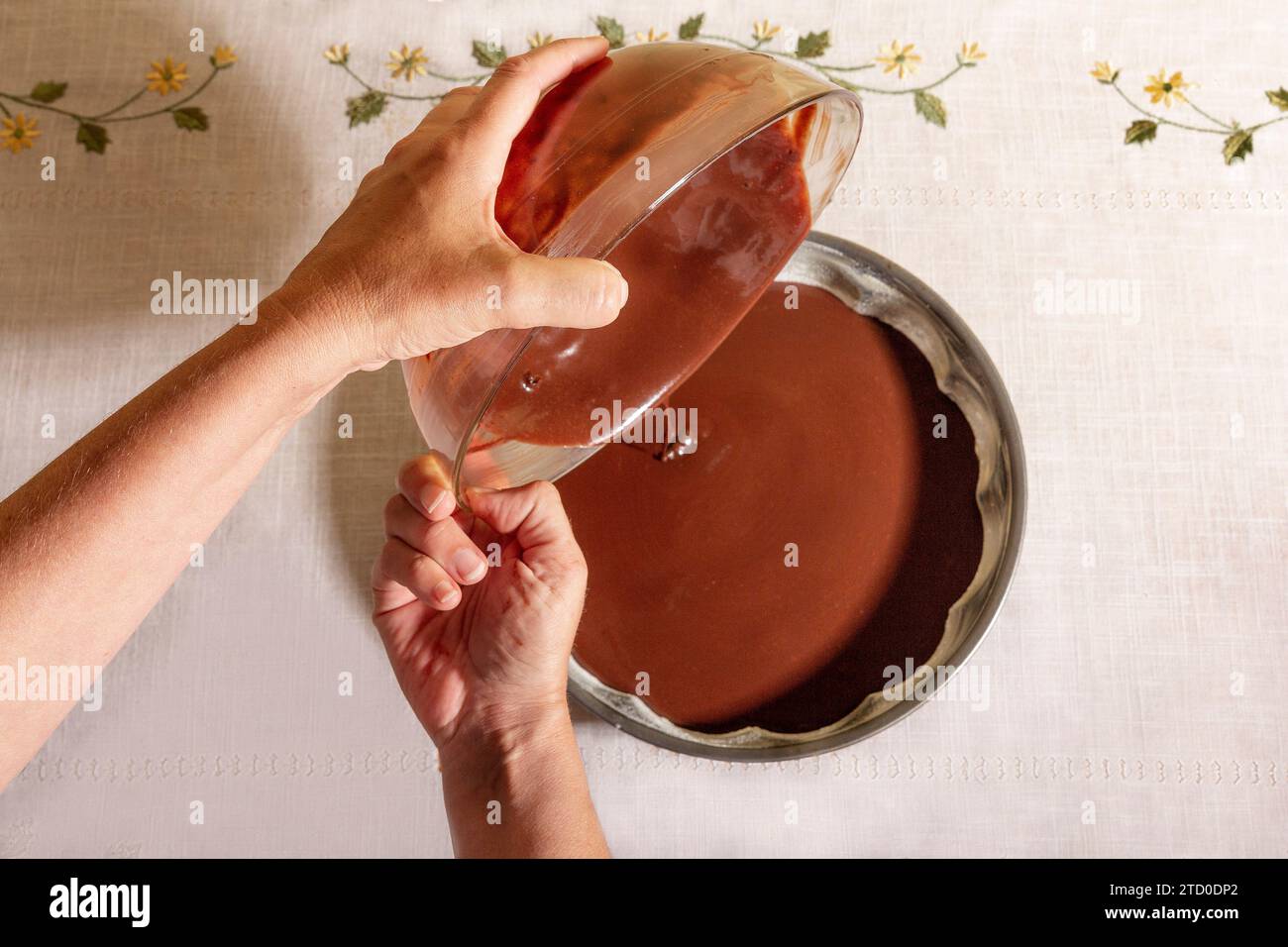 Vue de dessus des mains d'une personne anonyme versant soigneusement la pâte à chocolat lisse d'un bol en verre dans une plaque de cuisson ronde sur une table Banque D'Images