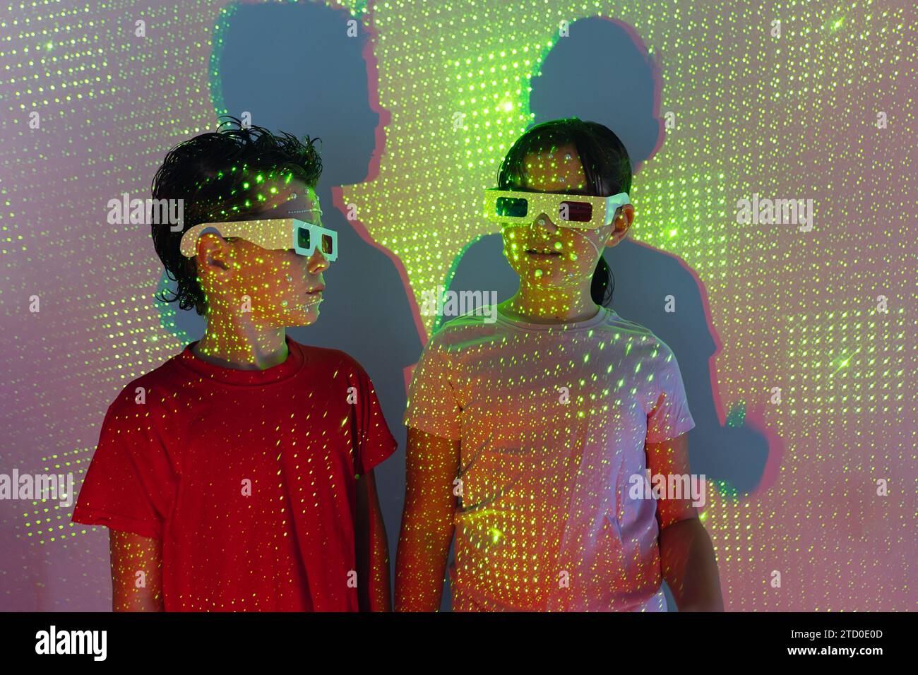 Frère et sœur positifs dans des vêtements décontractés portant des lunettes 3D debout ensemble contre le mur blanc dans des lumières au néon Banque D'Images