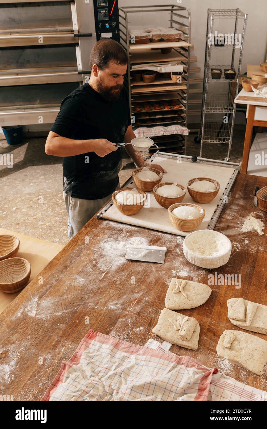 Vue à angle élevé de boulanger mâle focalisé tamisant la farine sur la pâte crue dans des bols à la table en bois tout en préparant la pâtisserie fraîche dans la cuisine à la boulangerie Banque D'Images