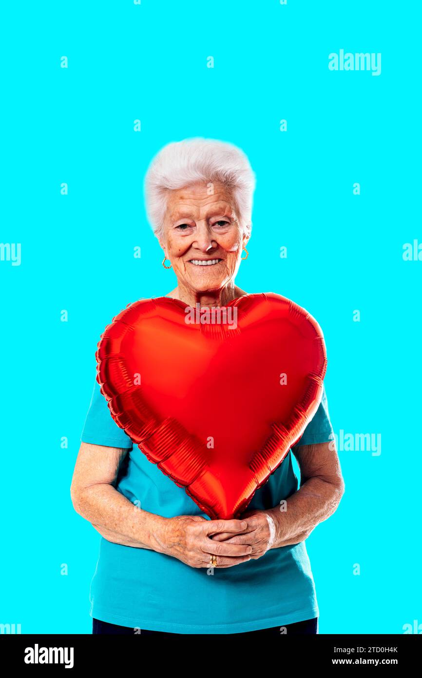 Femme âgée positive aux cheveux gris tenant un ballon en forme de coeur rouge et regardant la caméra sur fond bleu Banque D'Images
