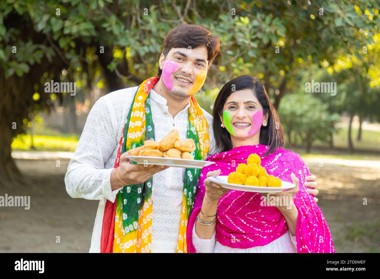 Heureux Jeune couple indien portant kurta blanc tenant une assiette pleine de Gujiyas et de ladoos faits maison. Célébration du festival Holi dans le parc ou le jardin. Banque D'Images