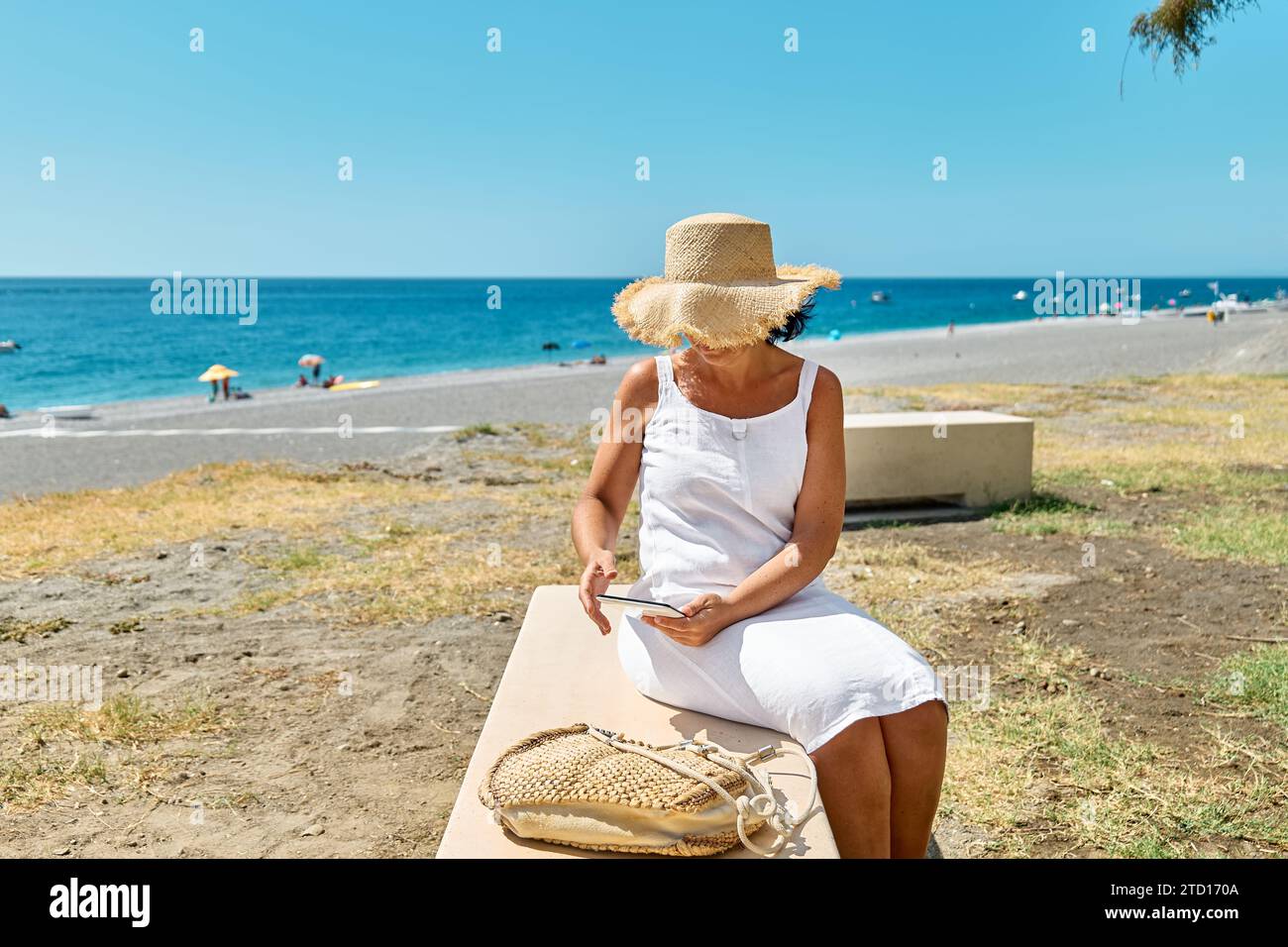 Femme mature portant un chapeau de paille en utilisant une tablette numérique à la plage. Femme d'âge moyen utilisant un appareil numérique pour faire des achats en ligne, des banques, des médias sociaux o Banque D'Images