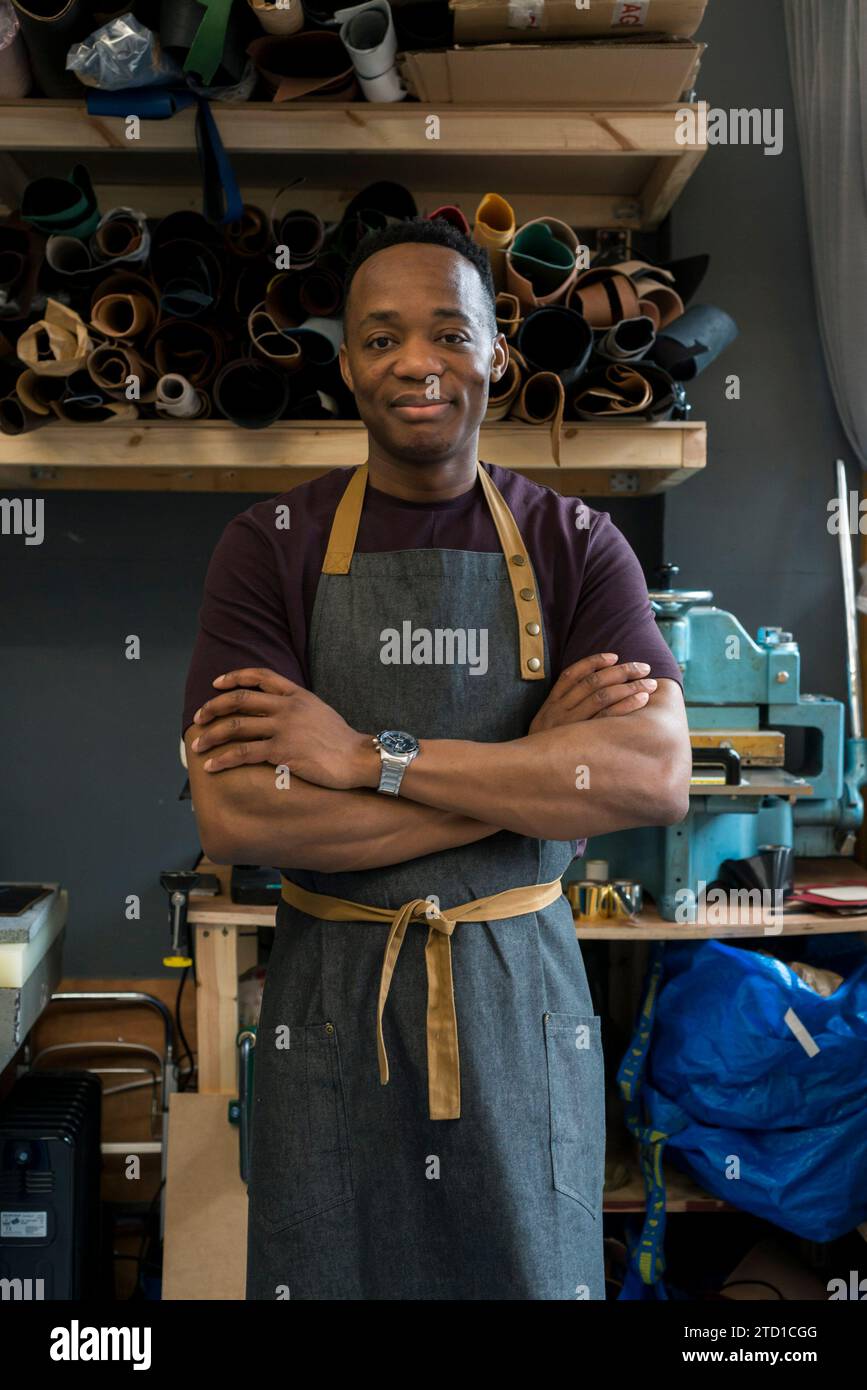 Un jeune homme se tient debout dans son atelier de cuir où il conçoit et fabrique des articles de mode tels que des sacs suspendus, des portefeuilles et des ceintures. Banque D'Images