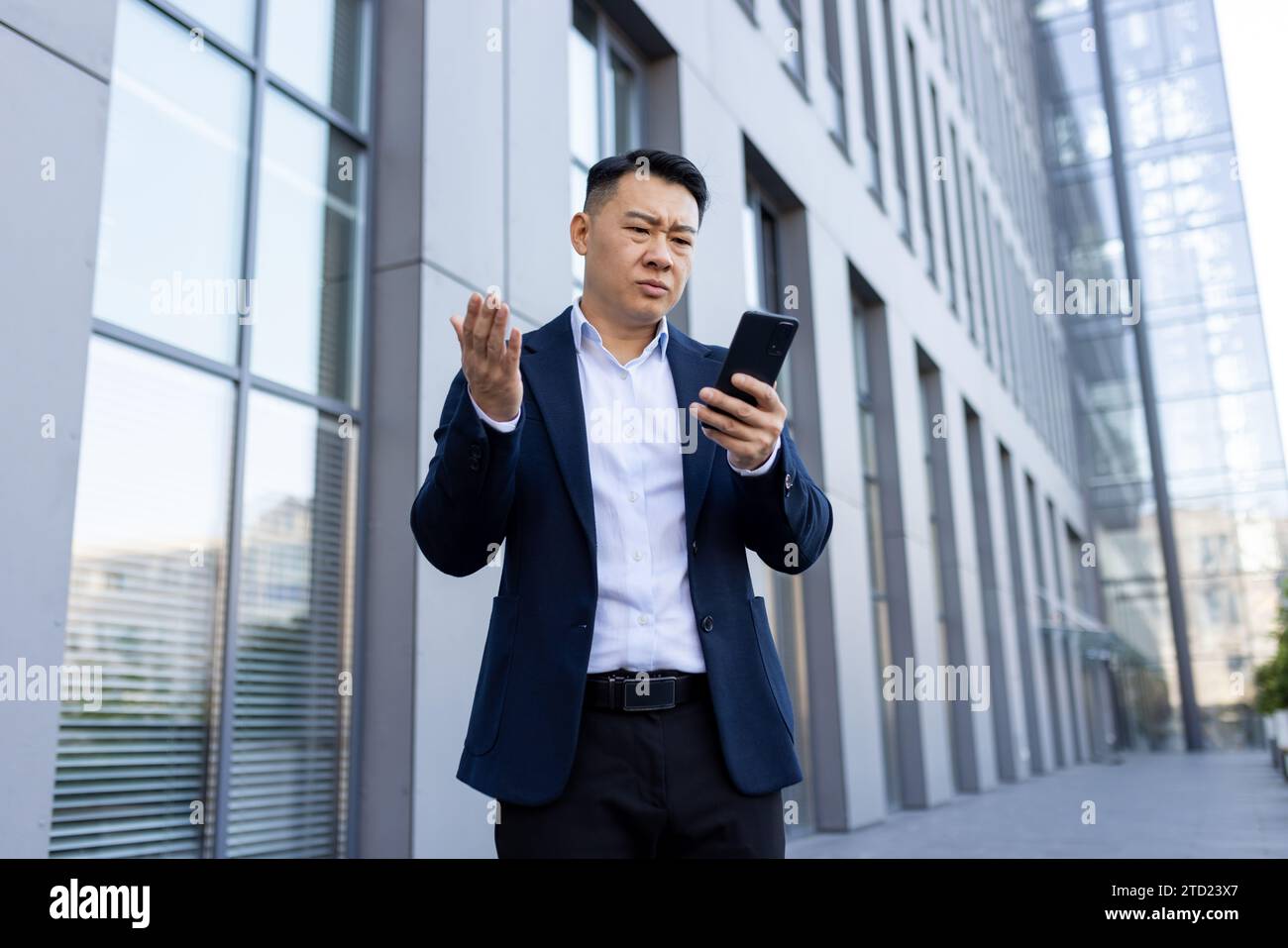 Jeune homme d'affaires asiatique sérieux debout à l'extérieur de l'immeuble de bureaux, tenant le téléphone, semblant frustré et inquiet à l'écran avec les bras tendus. Banque D'Images