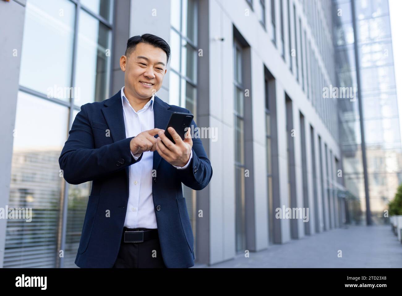 Un jeune homme d'affaires asiatique souriant en costume se tient devant un centre de bureau et utilise un téléphone portable. Banque D'Images