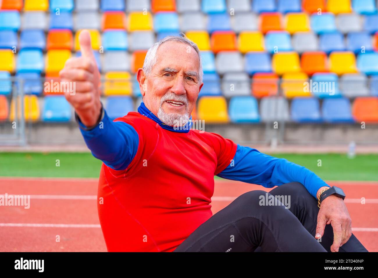 Homme âgé fier faisant des gestes avec les pouces vers le haut assis sur une piste de course après avoir couru Banque D'Images
