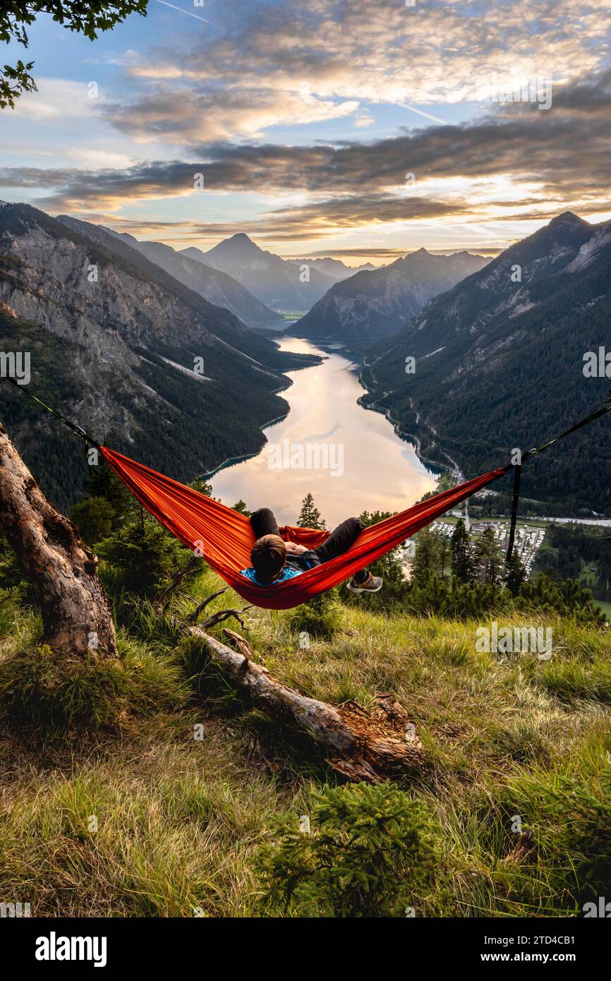 Jeune homme assis dans un hamac orange, vue de Plansee de Schoenjoechl, au coucher du soleil, vue panoramique sur les montagnes avec lac, Plansee, Tyrol, Autriche Banque D'Images