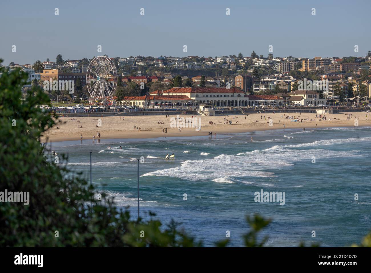 L'emblématique Bondi Beach et Bondi Pavilion, à Sydney, en Australie. Les gens dans le sable, l'eau et portant des planches de surf. Arbustes verts au premier plan. Banque D'Images